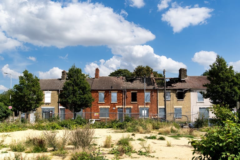 Derelict brick row houses with boarded windows surrounded by overgrown vegetation and sandy ground under a cloudy blue sky